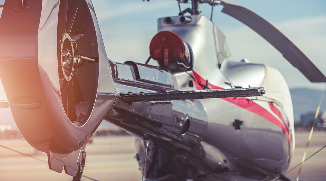 Close-up of a polished silver helicopter on a runway, showing the tail rotor and red accent stripe, sunlight reflecting off the fuselage.