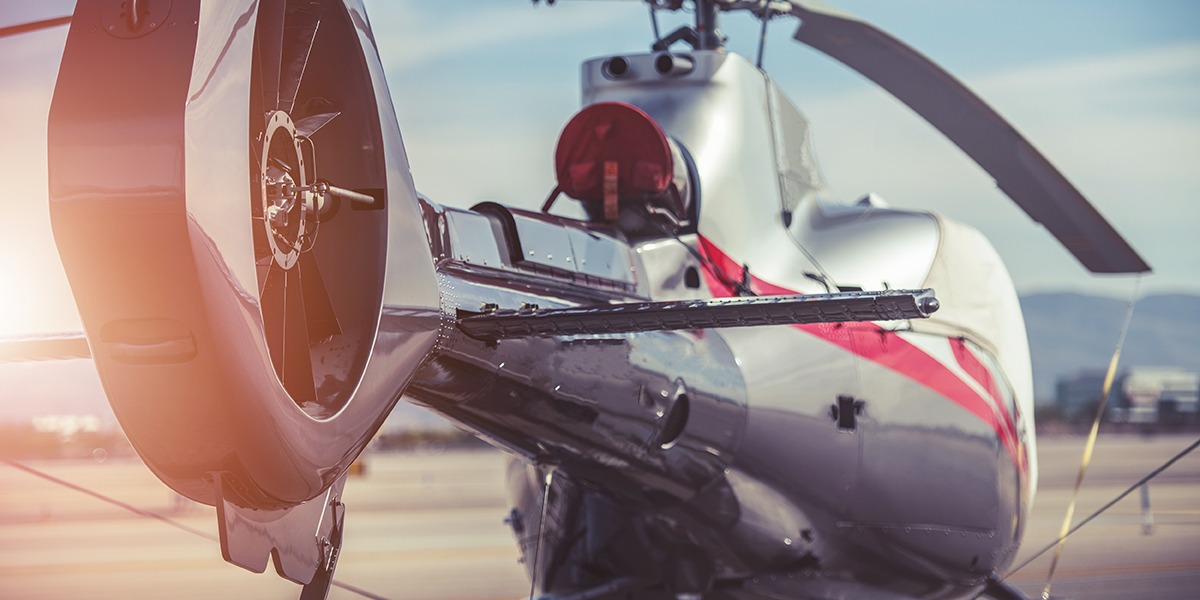 Close-up of a polished silver helicopter on a runway, showing the tail rotor and red accent stripe, sunlight reflecting off the fuselage.